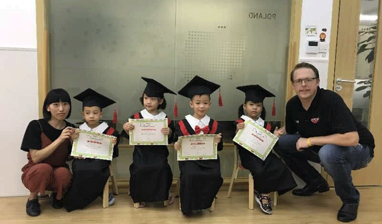 Man squatting with young Chinese students in graduation gowns and caps with fellow teacher