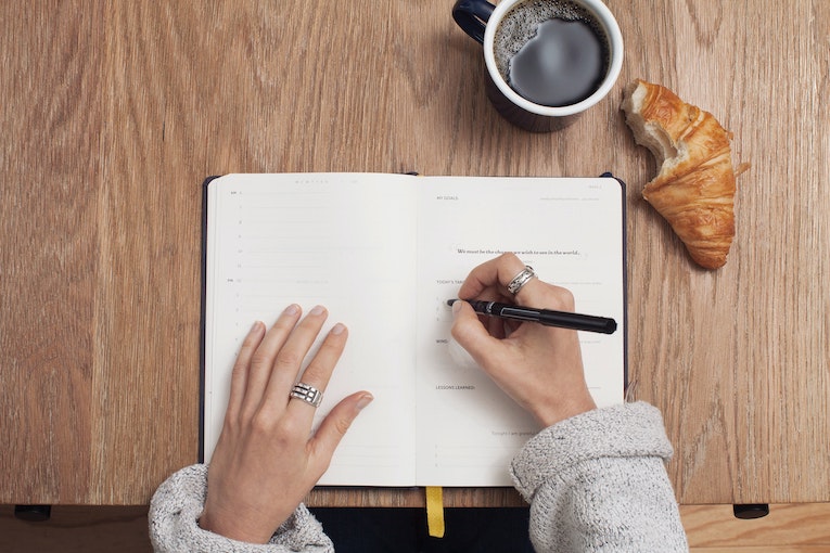 hands checking things off in a notebook with coffee and croissant to the side
