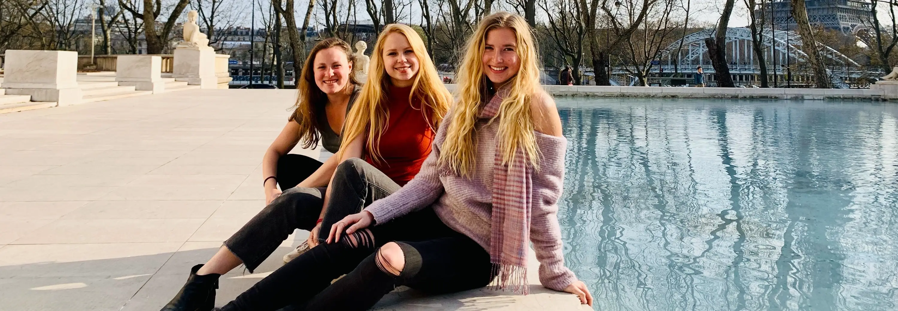 Three women smiling by a pool with Eiffel Tower in the background.