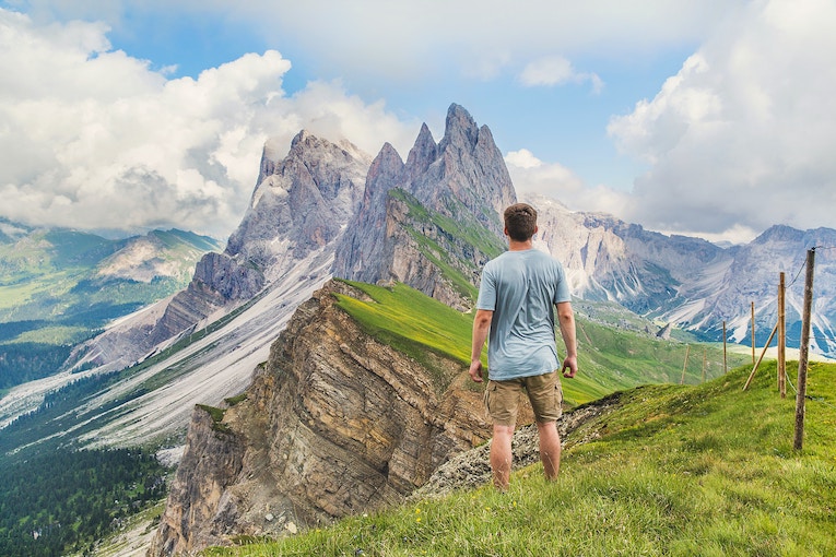 person in T-shirt and shorts at top of a mountain looking at higher mountains in distance