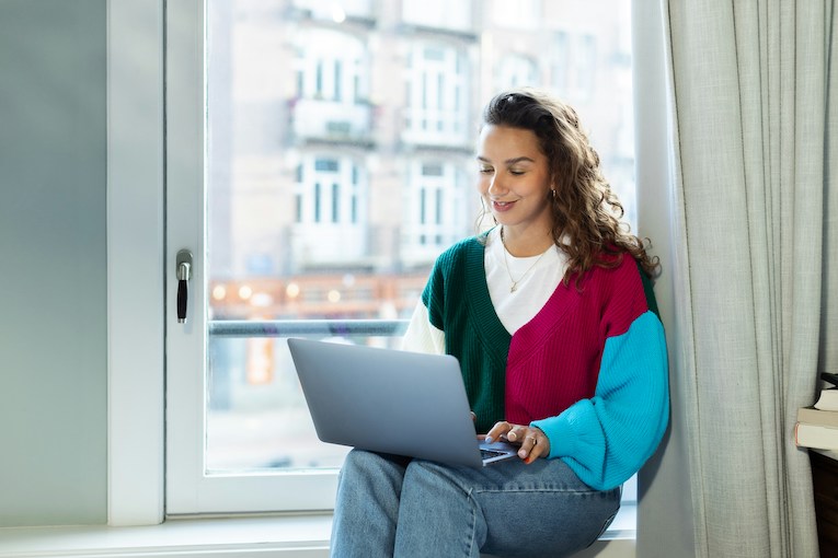 smiling person sitting on windowsill using laptop
