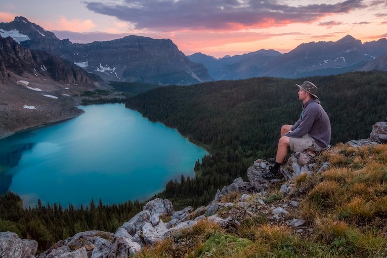 person sitting on rock looking at body of water and mountains under colorful sky