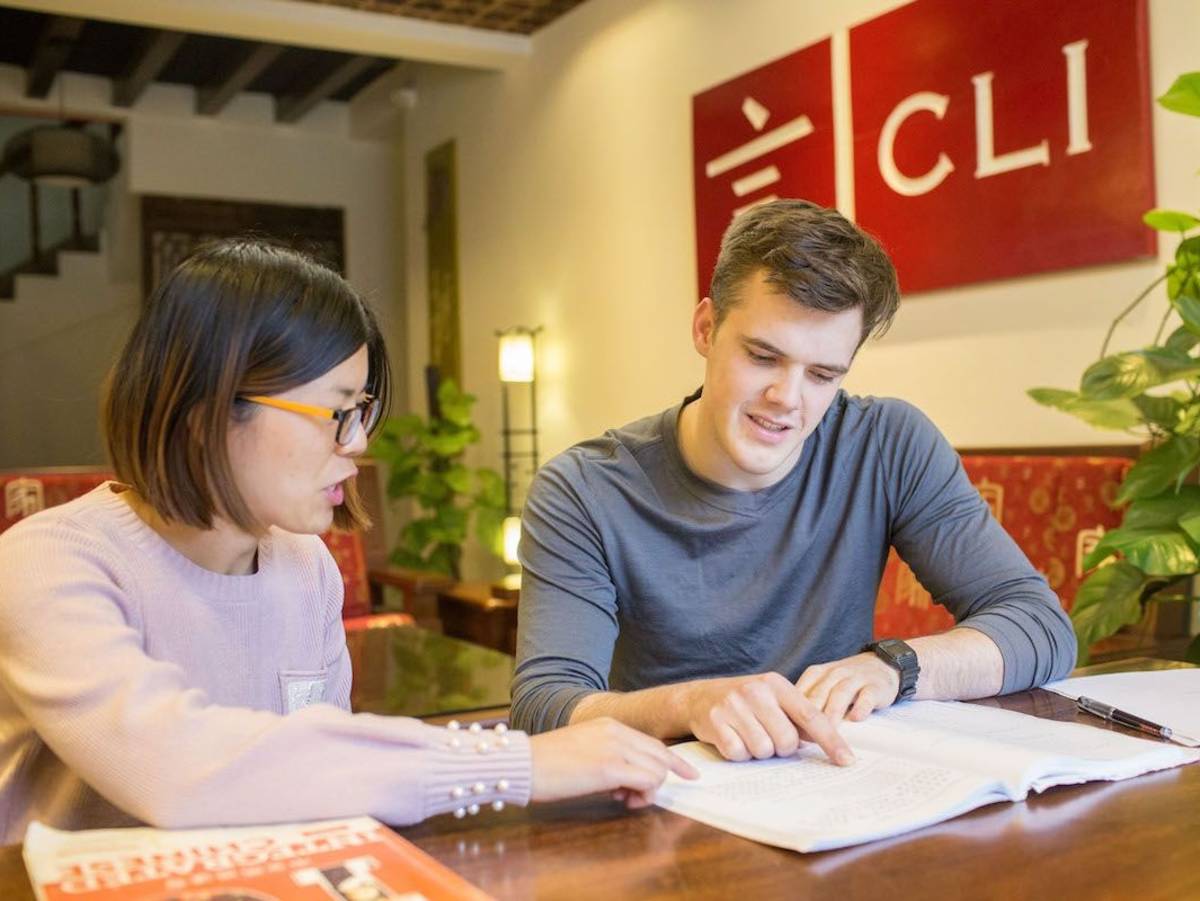 A CLI teacher and student review a Chinese textbook together at a wooden table, with the red CLI logo on the wall behind them.