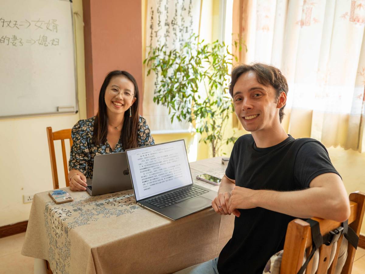 A CLI teacher and student smile at the camera during a one-on-one Chinese lesson, with laptops open and Chinese vocabulary written on a whiteboard behind them.