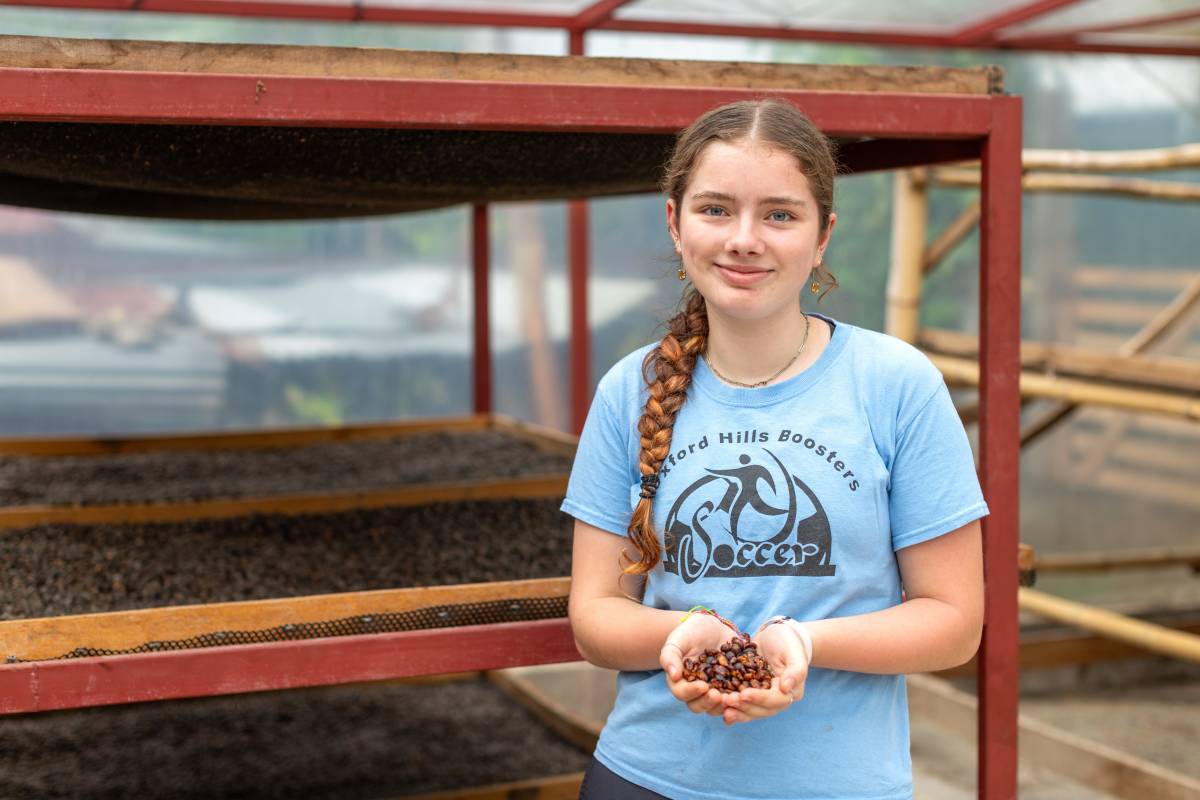 A girl poses for the camera, holding coffee beans cupped in her hand with drying racks in the background.