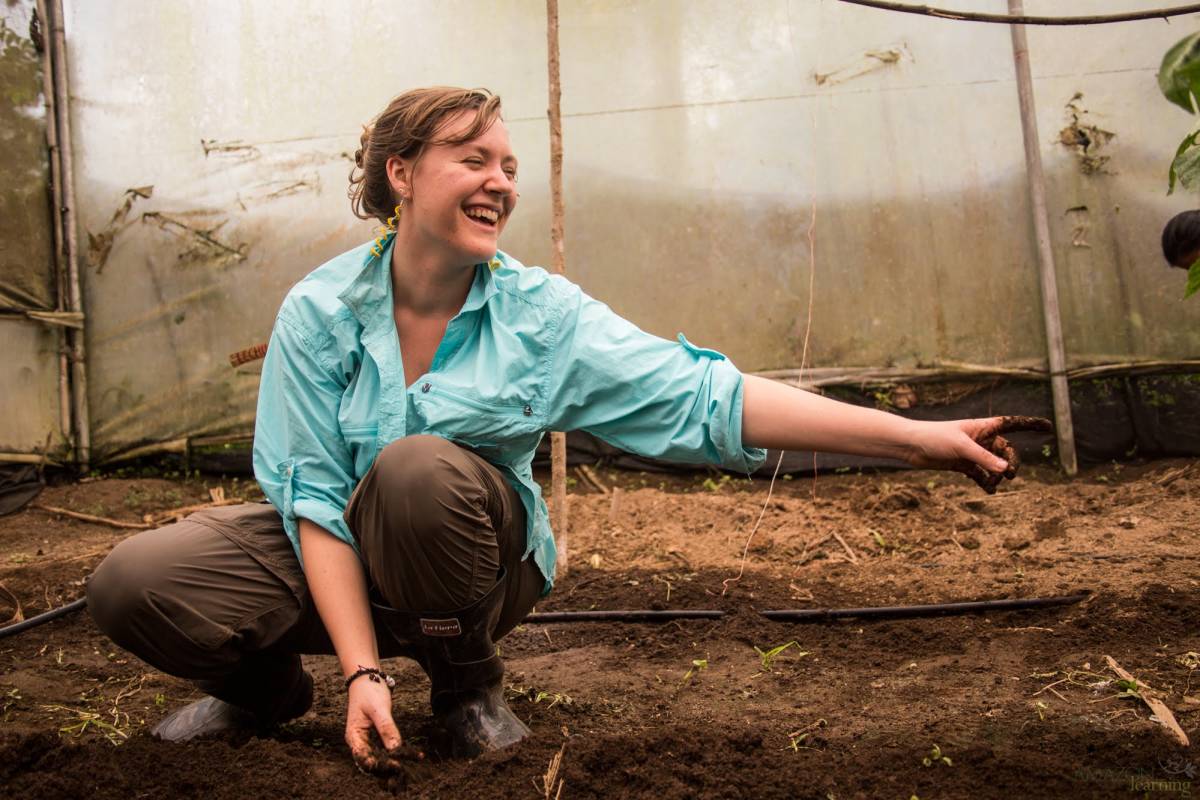 A girl smiles and looks to the side as she is crouched in greenhouse