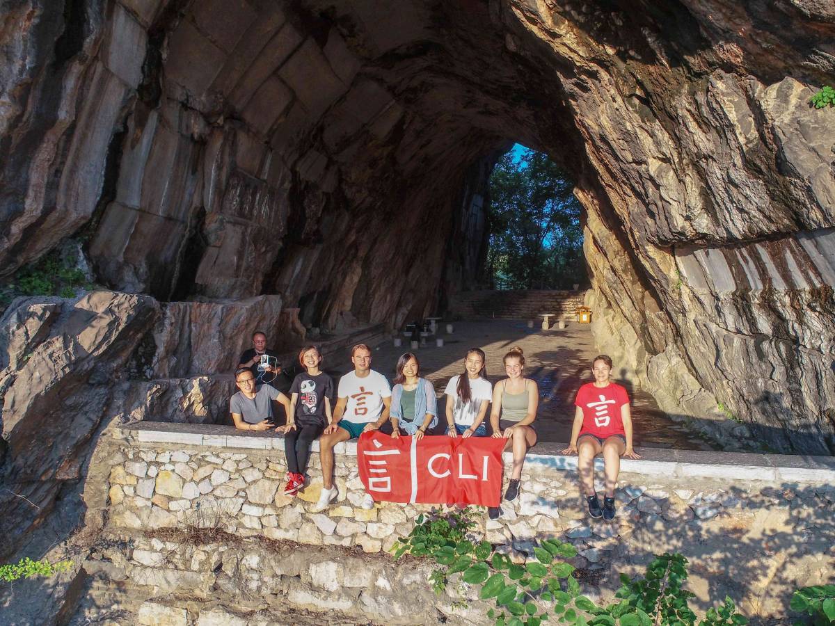 A group of CLI students and teachers sit on a stone wall holding a red CLI flag at the entrance of a large karst cave in Guilin.
