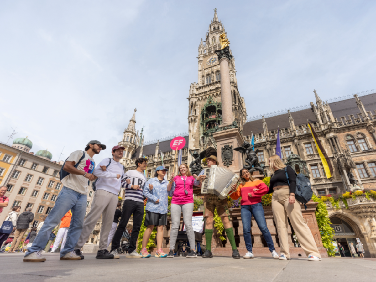 A group of EF Language Abroad students with a staff member stand in Marienplatz, Munich, near a street performer playing accordion in front of the Neues Rathaus.