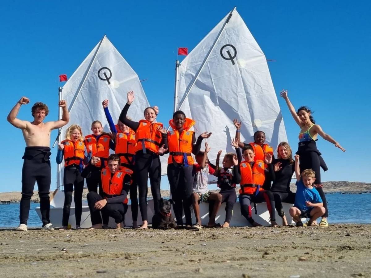 A group of interns and students in wetsuits and orange life jackets pose cheerfully on the beach in front of two white sailing dinghies.