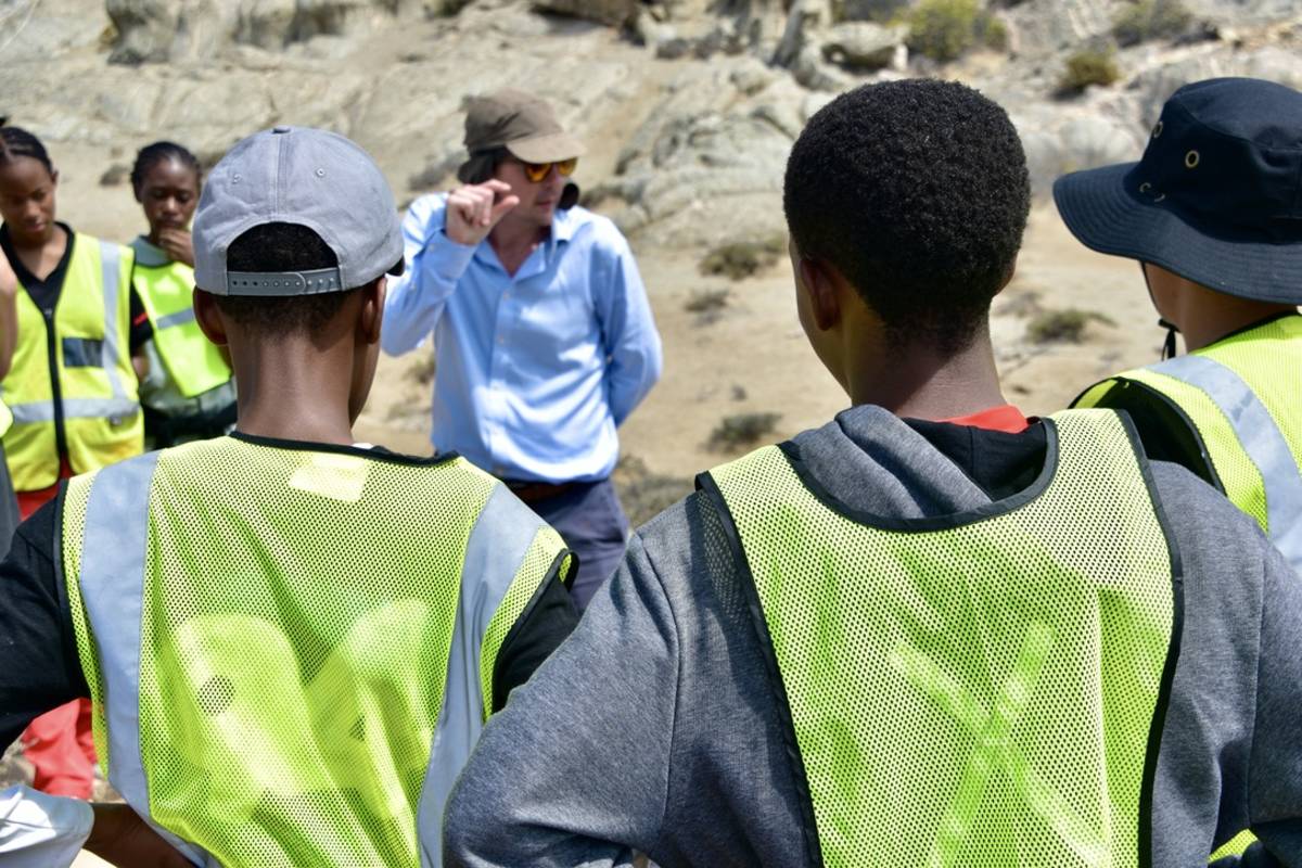 A group of Lüderitz Blue School students in high-visibility vests listening to an instructor teaching outdoors on the rocky Namibian coastline during an Ocean Day field session.