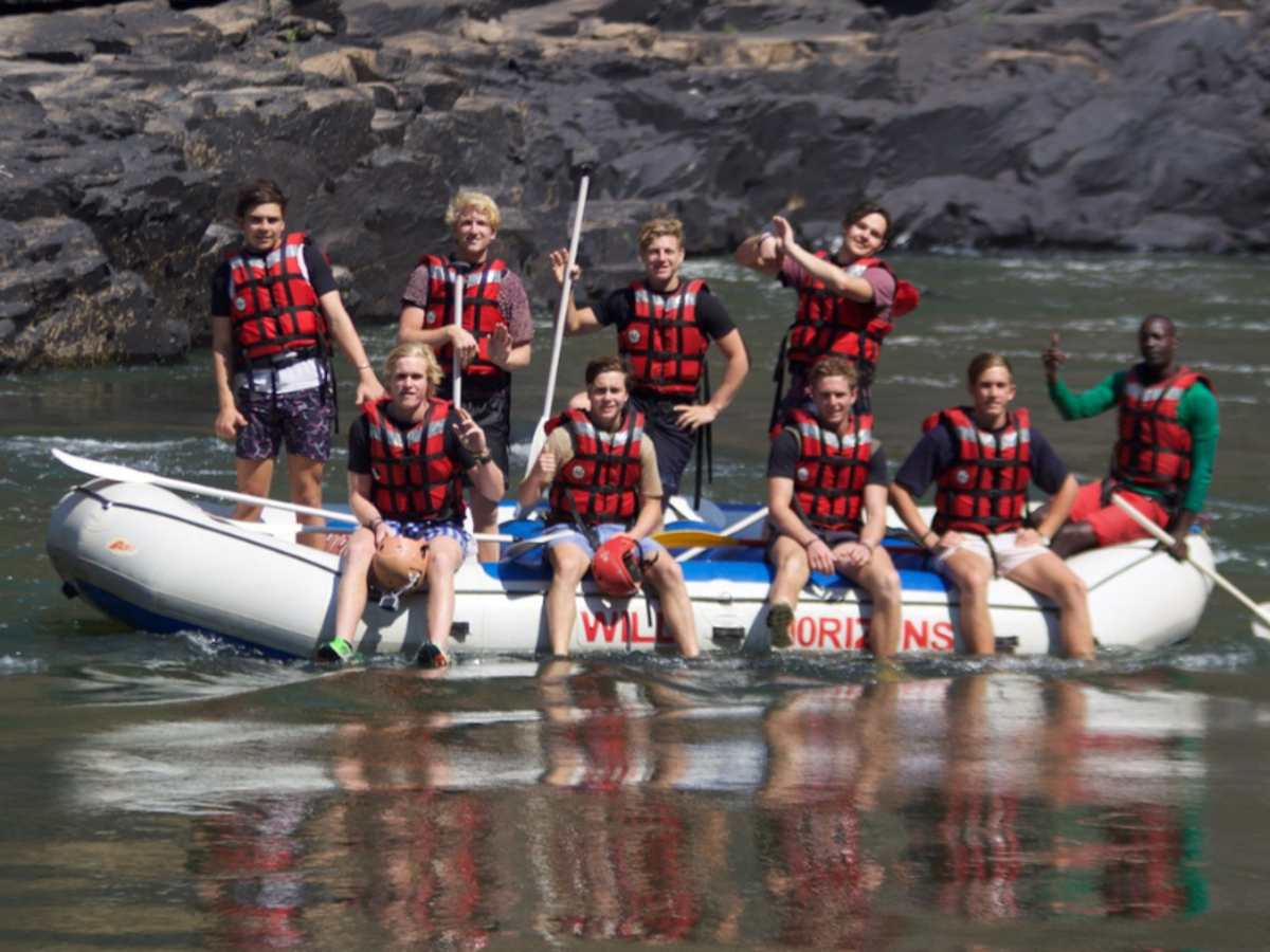 A group of nine people in life jackets pose on a raft in a river, smiling and making gestures. The scene conveys excitement and adventure, set against rocky banks.