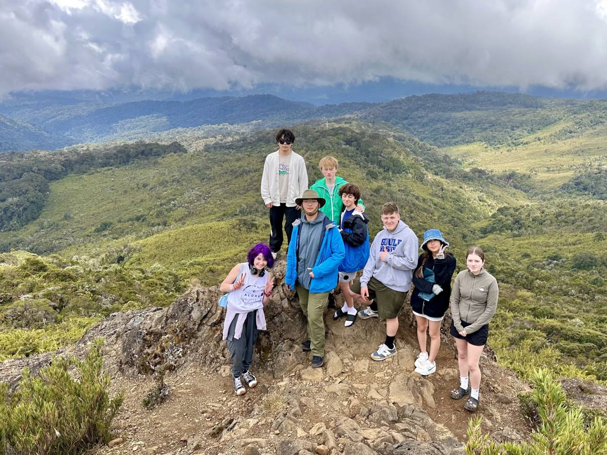 A group picture posed on top of a mountain, with green hills in background.