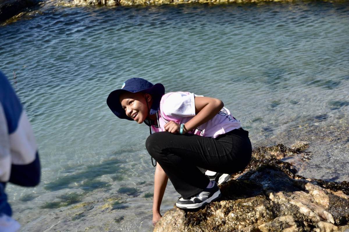 A Lüderitz Blue School boarding student crouching and smiling beside clear water at Diaz Point, Namibia.