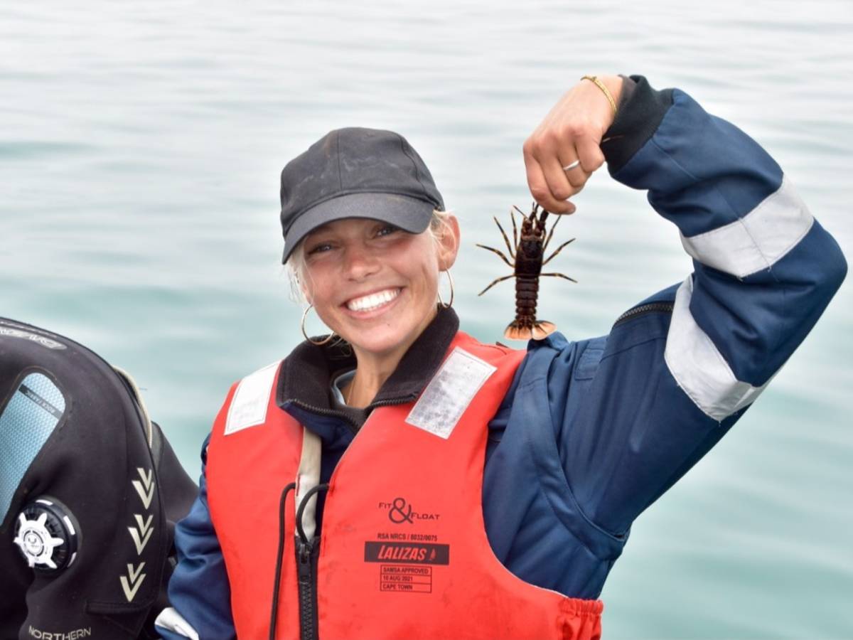 A smiling team member in a life jacket holds up a rock lobster retrieved during a crayfish monitoring survey on a research vessel off the Lüderitz coast.