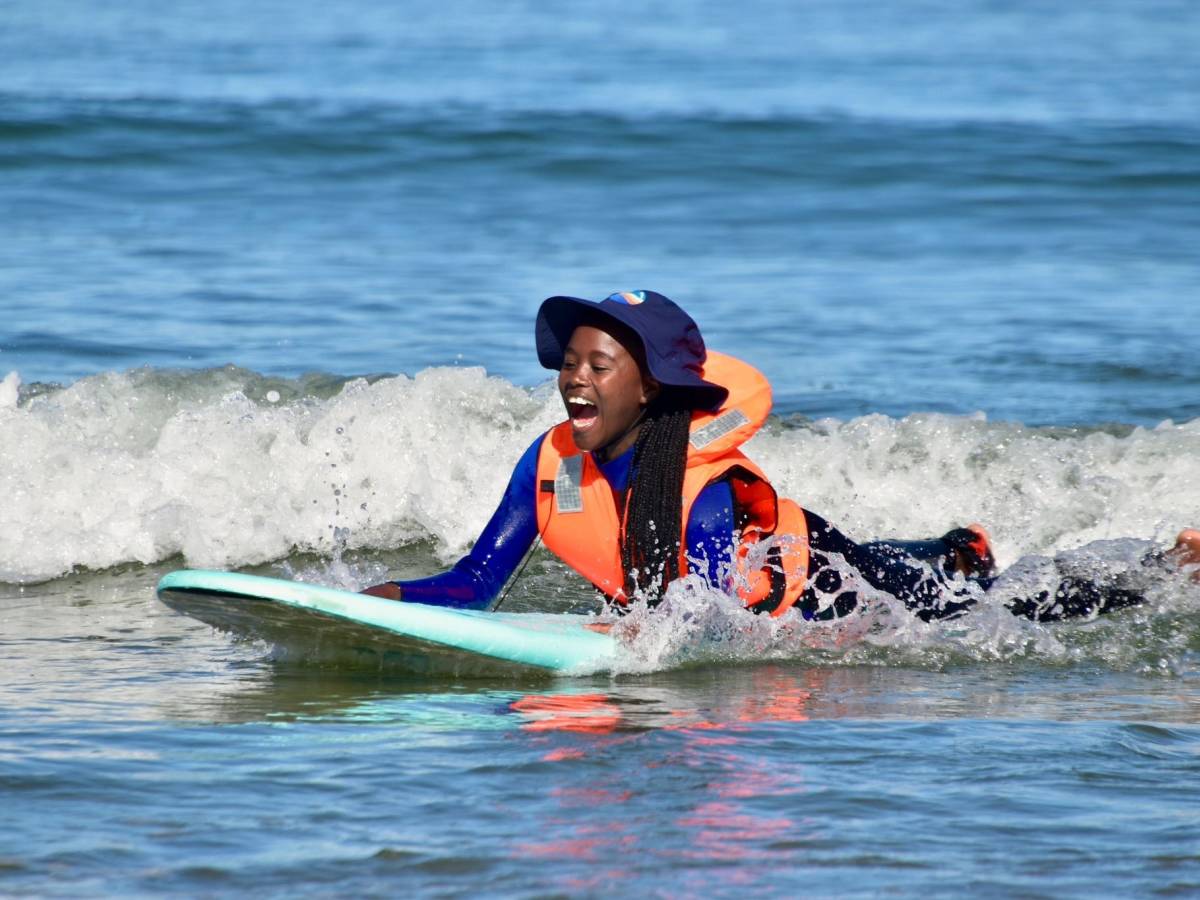 A student in an orange life jacket and hat laughs with joy as she rides a wave on a surfboard in the South Atlantic off the Lüderitz coast.