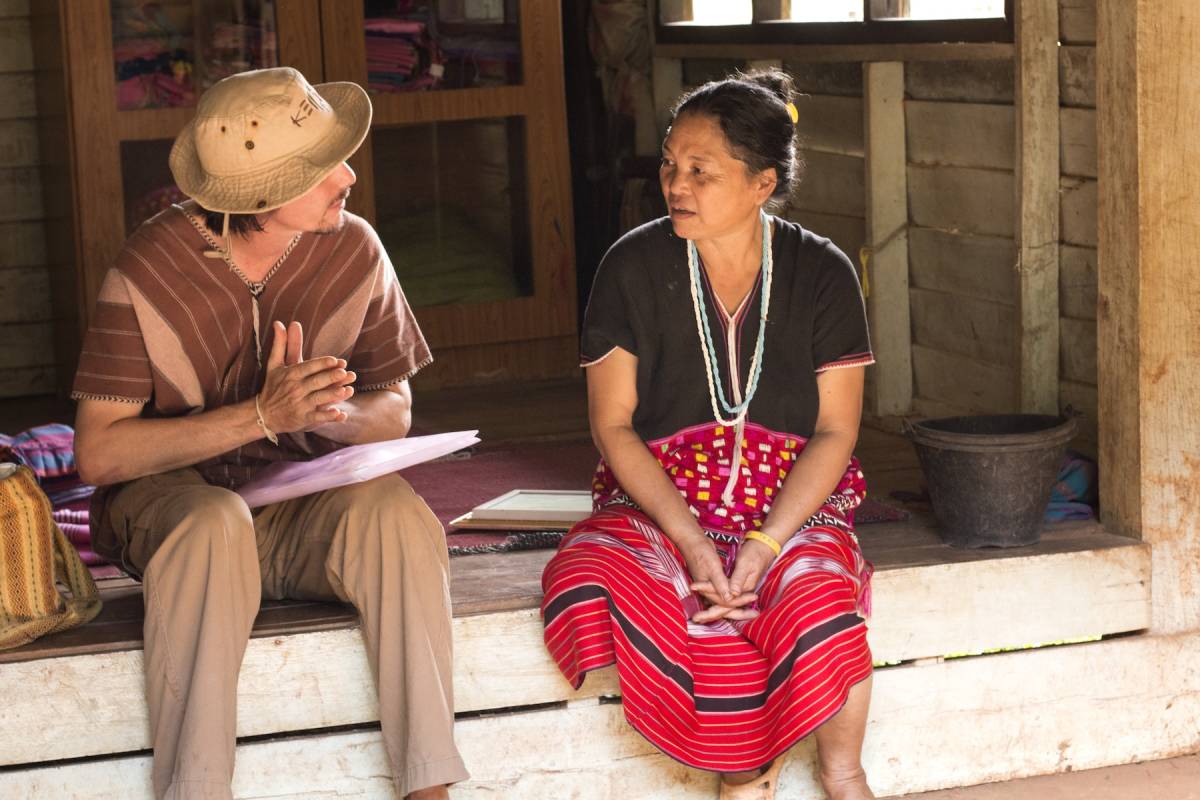 A student wearing a hat sits on a doorstep with a traditionally dressed woman and is asking her questions and writing in his notebook.