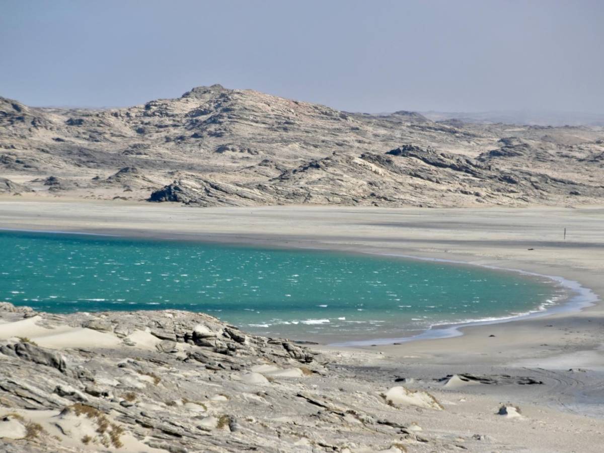 A turquoise lagoon nestled between dramatic rocky desert terrain and pale sand dunes on the Namibian coastline, under a hazy sky.