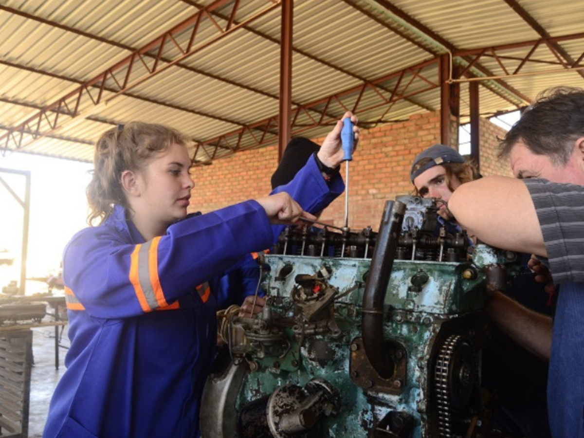 A woman in a blue work uniform with orange stripes repairs a large engine, surrounded by three men. The setting is industrial, with a focused atmosphere.