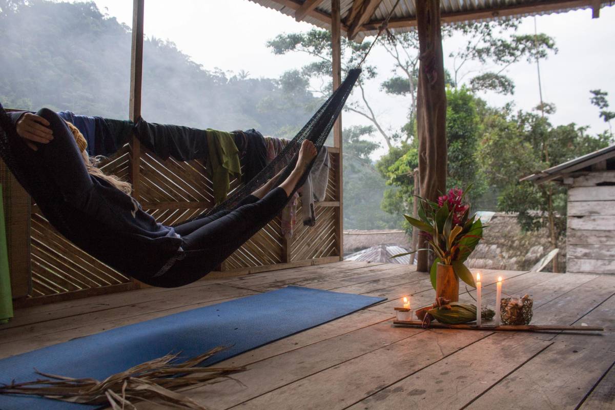 A woman relaxes in a hamock, looking off toward the river. Candles and flowers and a yoga mat are on the floor.