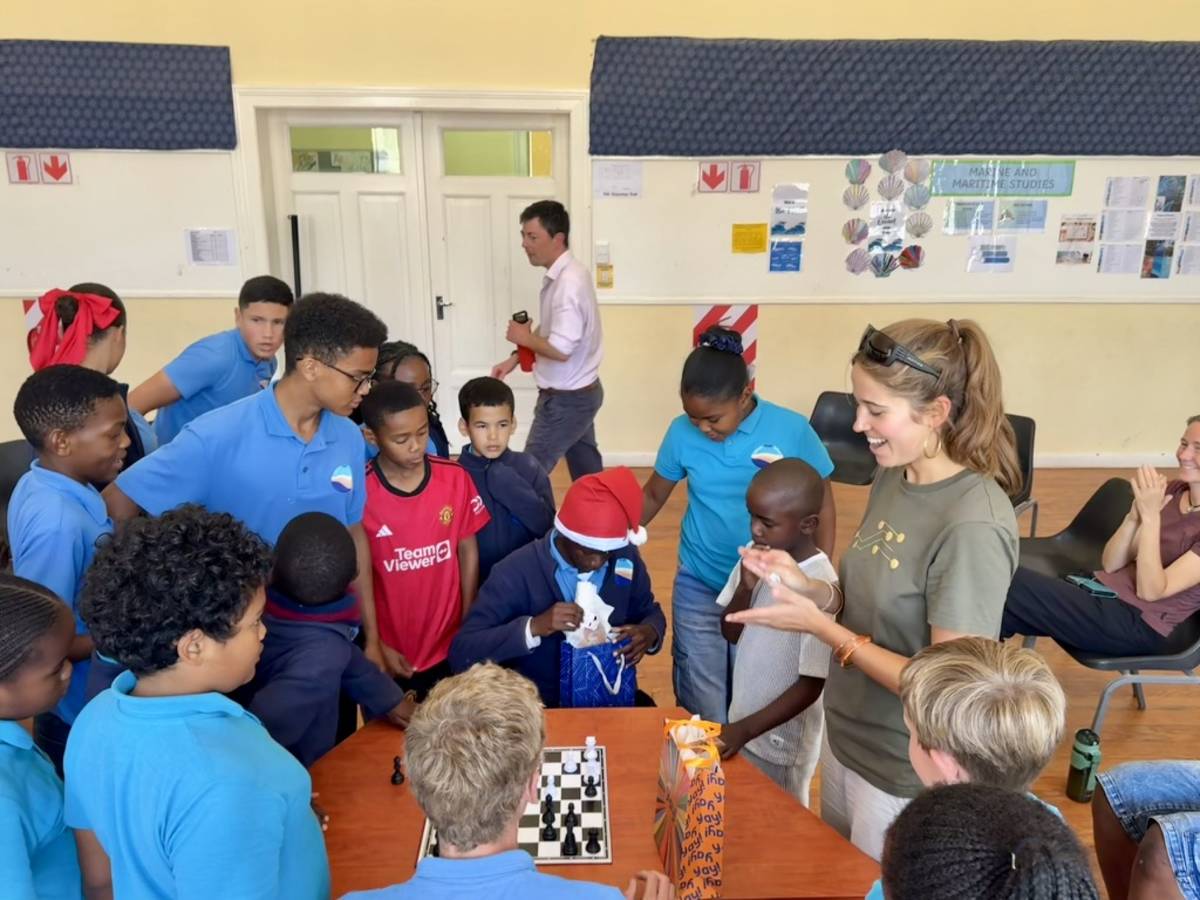 An intern and a group of primary school students crowd excitedly around a chess board during the final of a school chess knockout tournament.