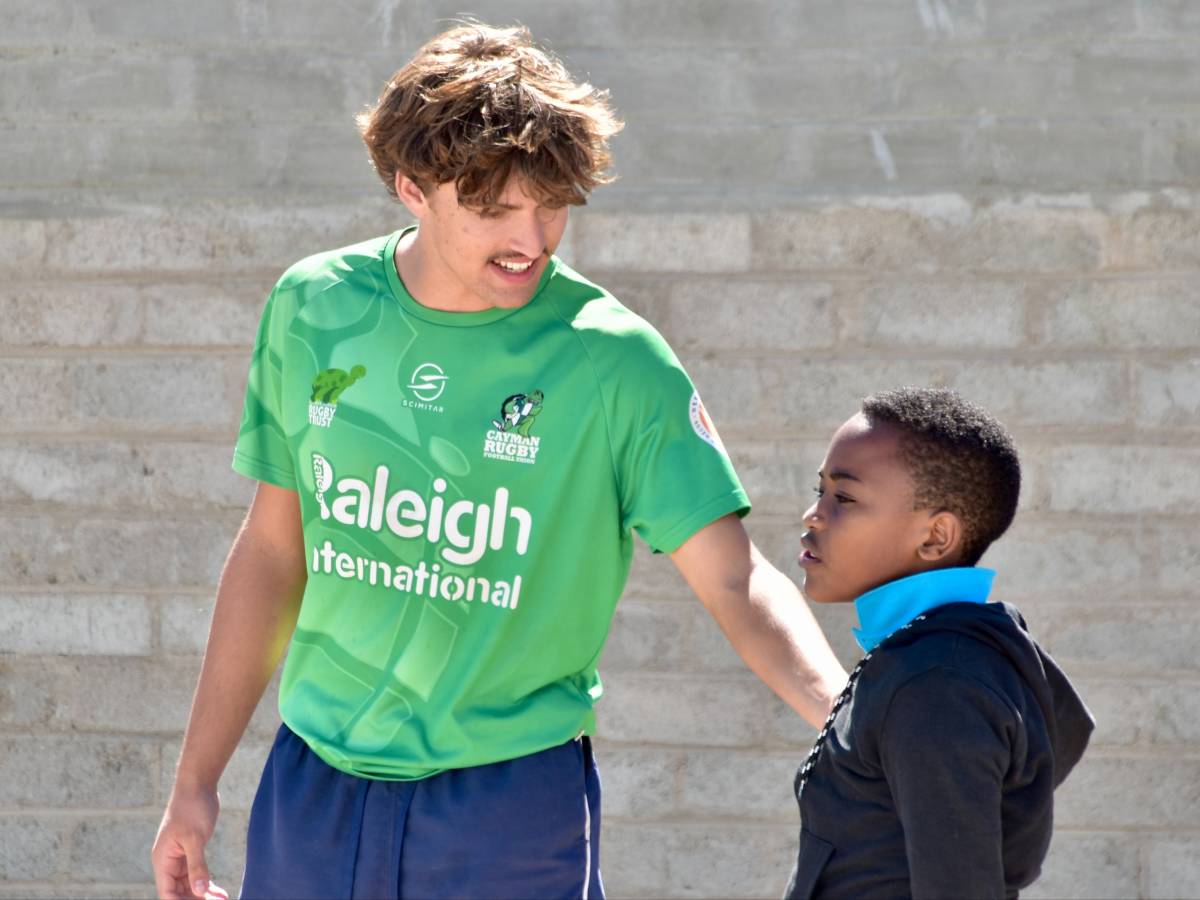 An intern in a green sports shirt crouches down to talk to a young primary school student on an outdoor sports court.