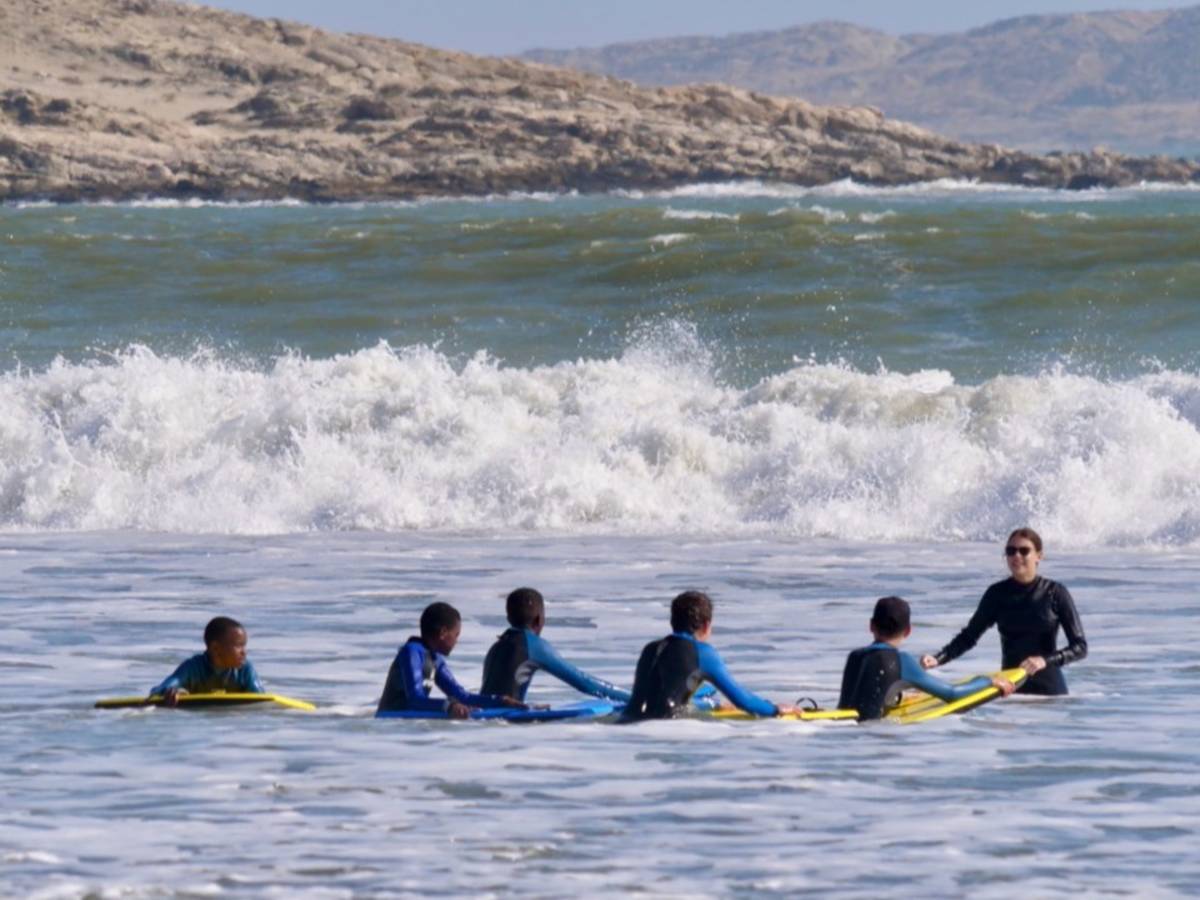 An intern in a wetsuit guides a line of primary school students on yellow bodyboards in the ocean, with crashing waves and the Lüderitz coastline behind them.