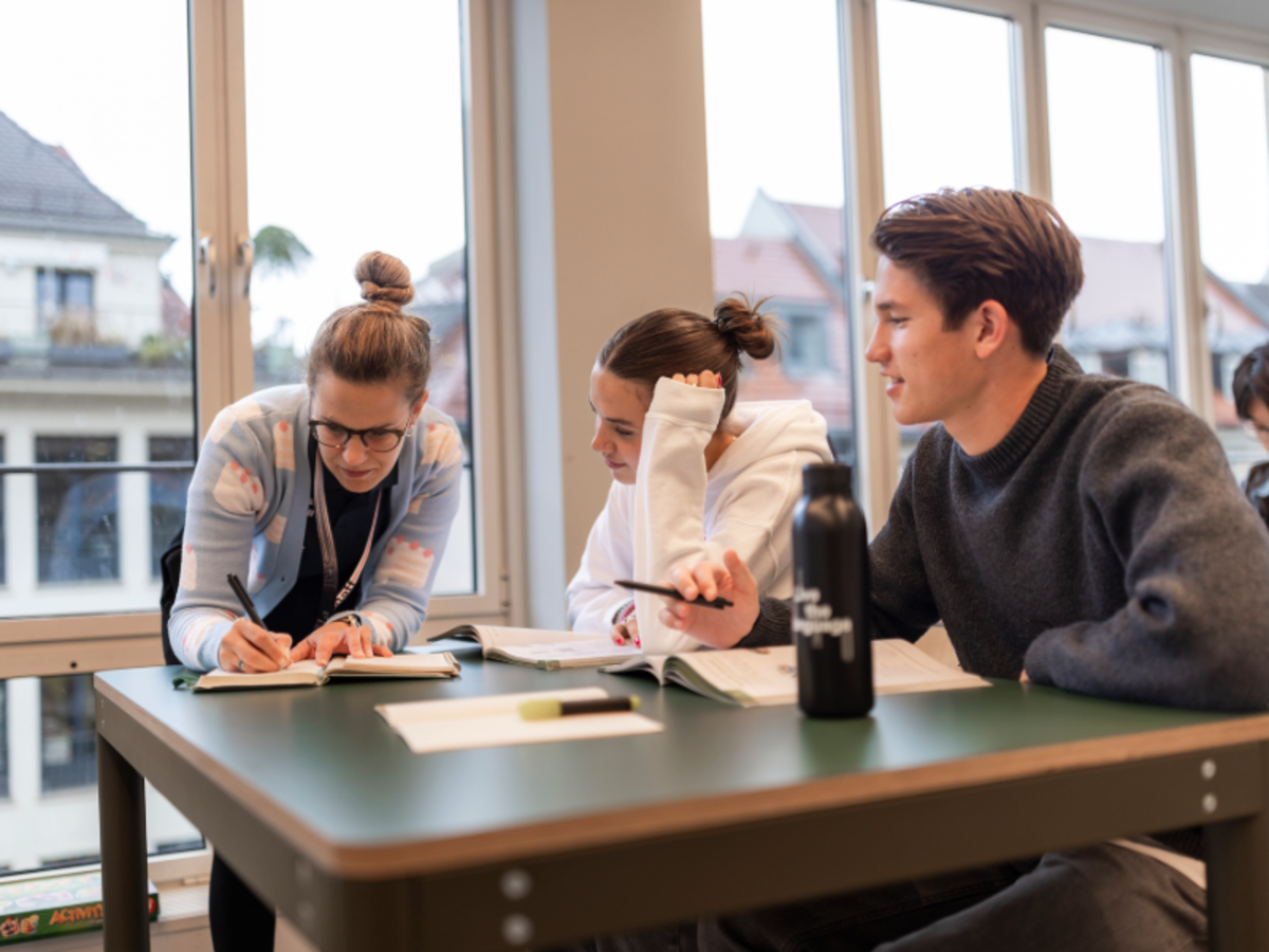 EF Language Abroad teacher helps two students with their German studies inside a bright Munich classroom with large windows.