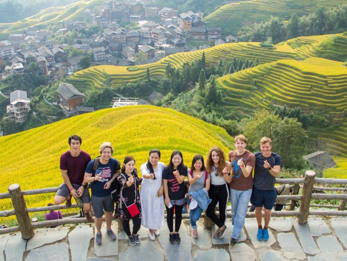 Nine students pose at a stone overlook above the Longji rice terraces, with terraced hillsides and a village behind them.