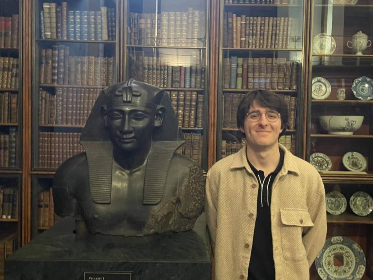 Person standing beside an ancient Egyptian statue in a library with bookshelves in the background.