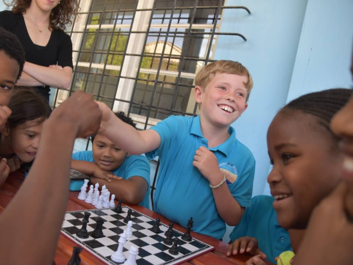 Primary school students react with excitement around a chess board during a knockout tournament game, with an intern supervising in the background.