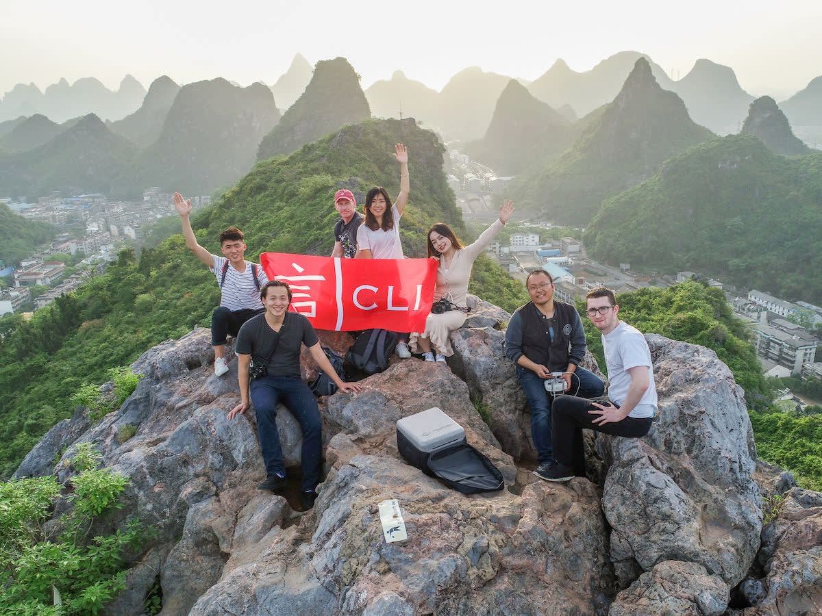 Seven CLI students and staff hold a red CLI banner on a rocky karst peak at sunset, with Guilin's mountains and city below.