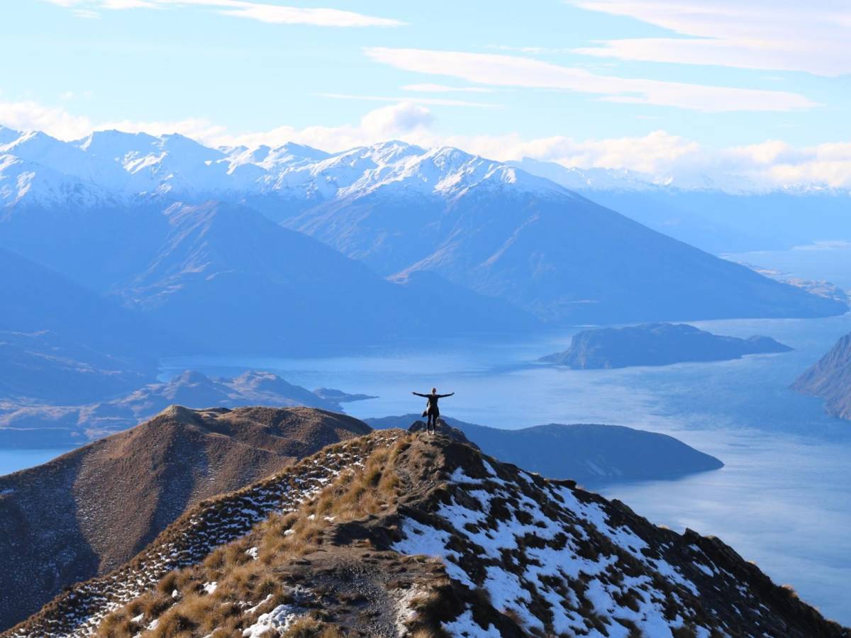 student standing on a mountain top in front of snowcapped mountains and a lake