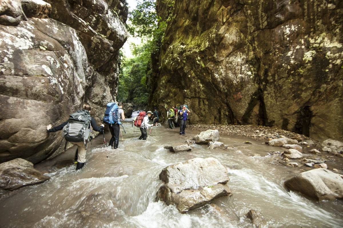 Students wearing backpacks, boots and carrying hiking poles traverse a river surrounding by rocky outcrops