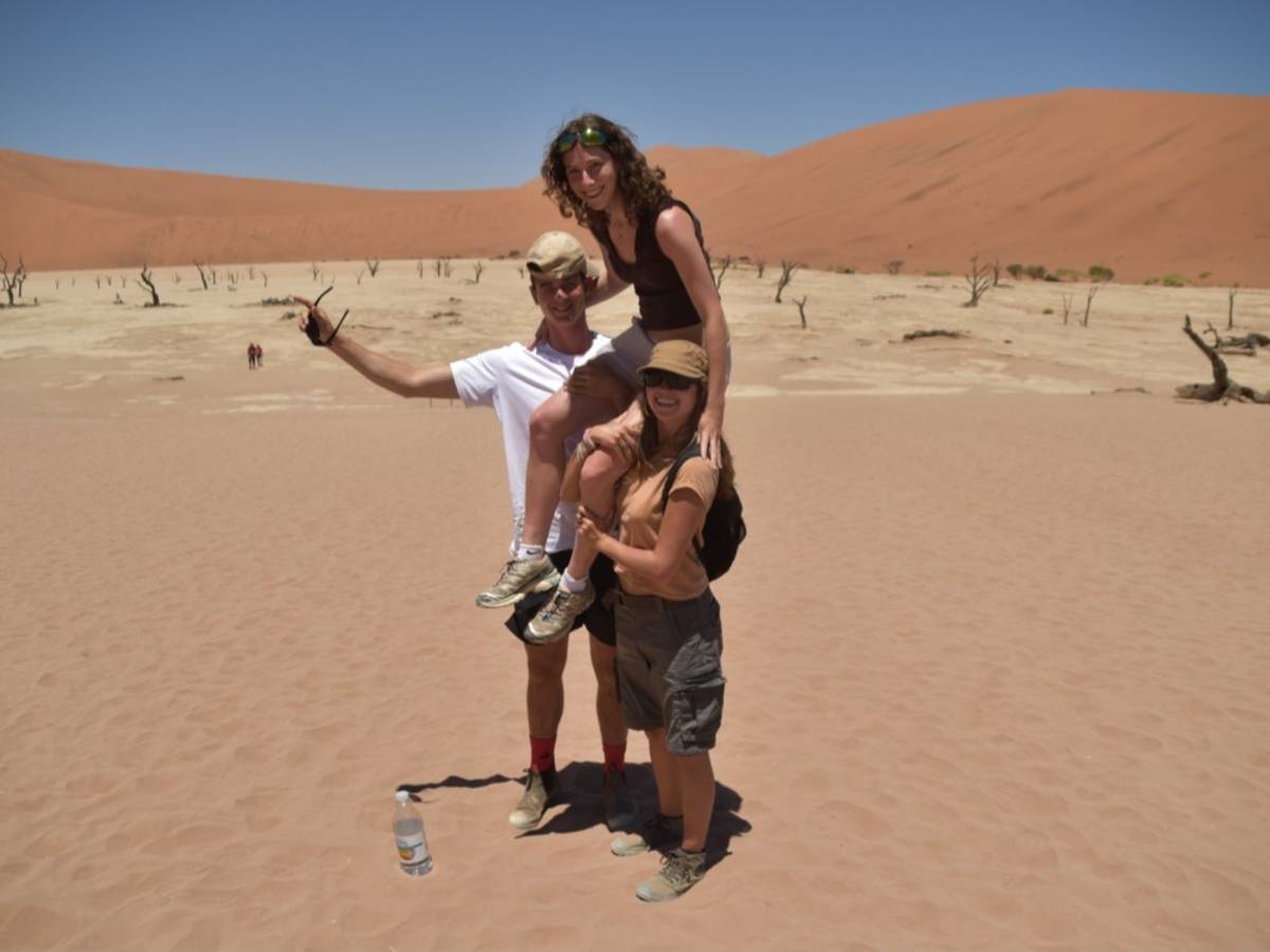 Three interns pose together at Deadvlei, surrounded by ancient dead trees and vast red sand dunes under a clear blue sky in the Namib Desert.