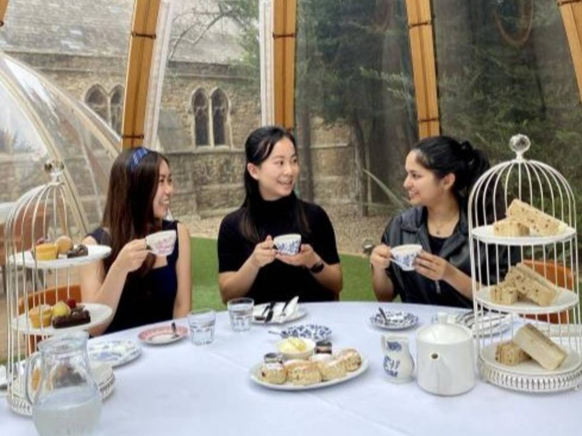 Three people seated at a table with tea cups, sandwiches, and pastries inside a glass conservatory.