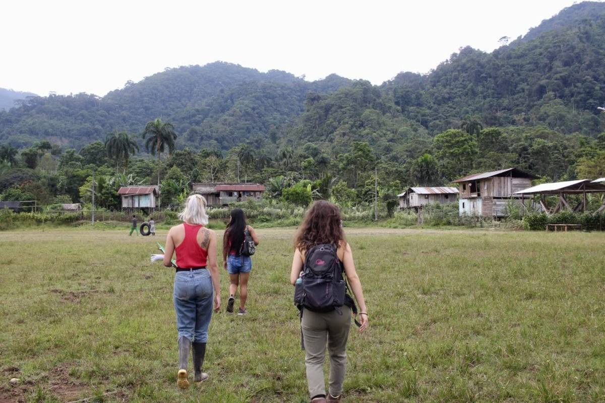Three women walk across a grassy field with houses and tropical mountains in background.