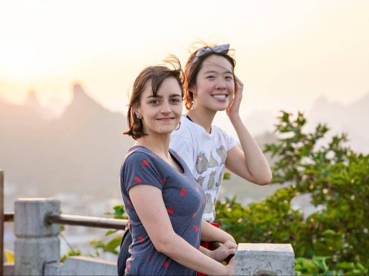 Two smiling students lean on a stone railing at a hilltop viewpoint, with hazy karst peaks and a sunset sky behind them.