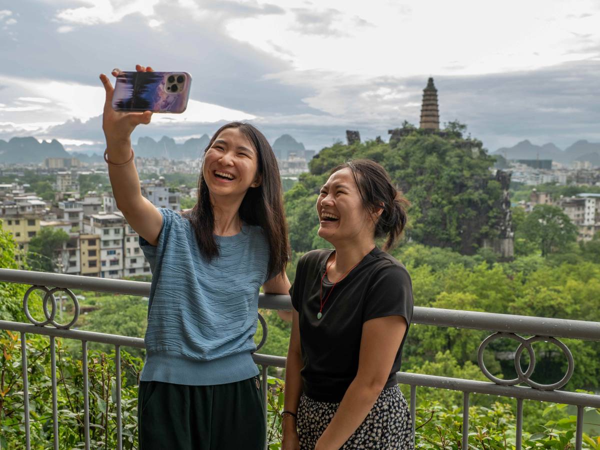 Two women laugh while taking a selfie at a hilltop viewpoint in Guilin, with a pagoda and karst mountains in the background.
