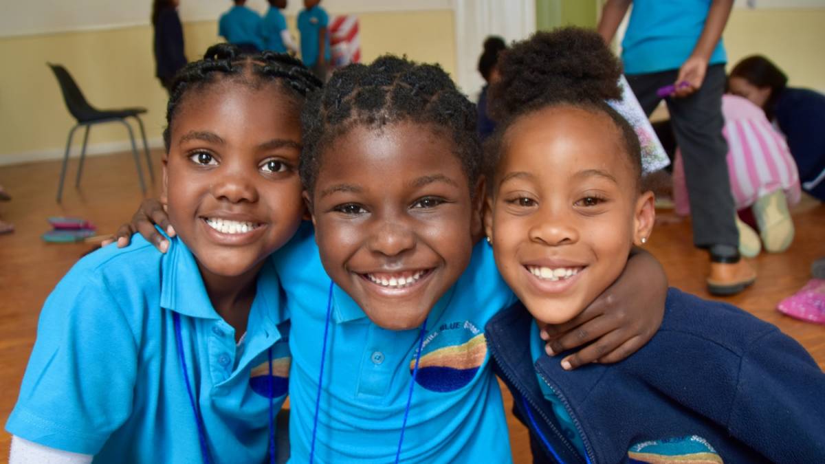 3 primary students in uniforms smiling with arms around each other.