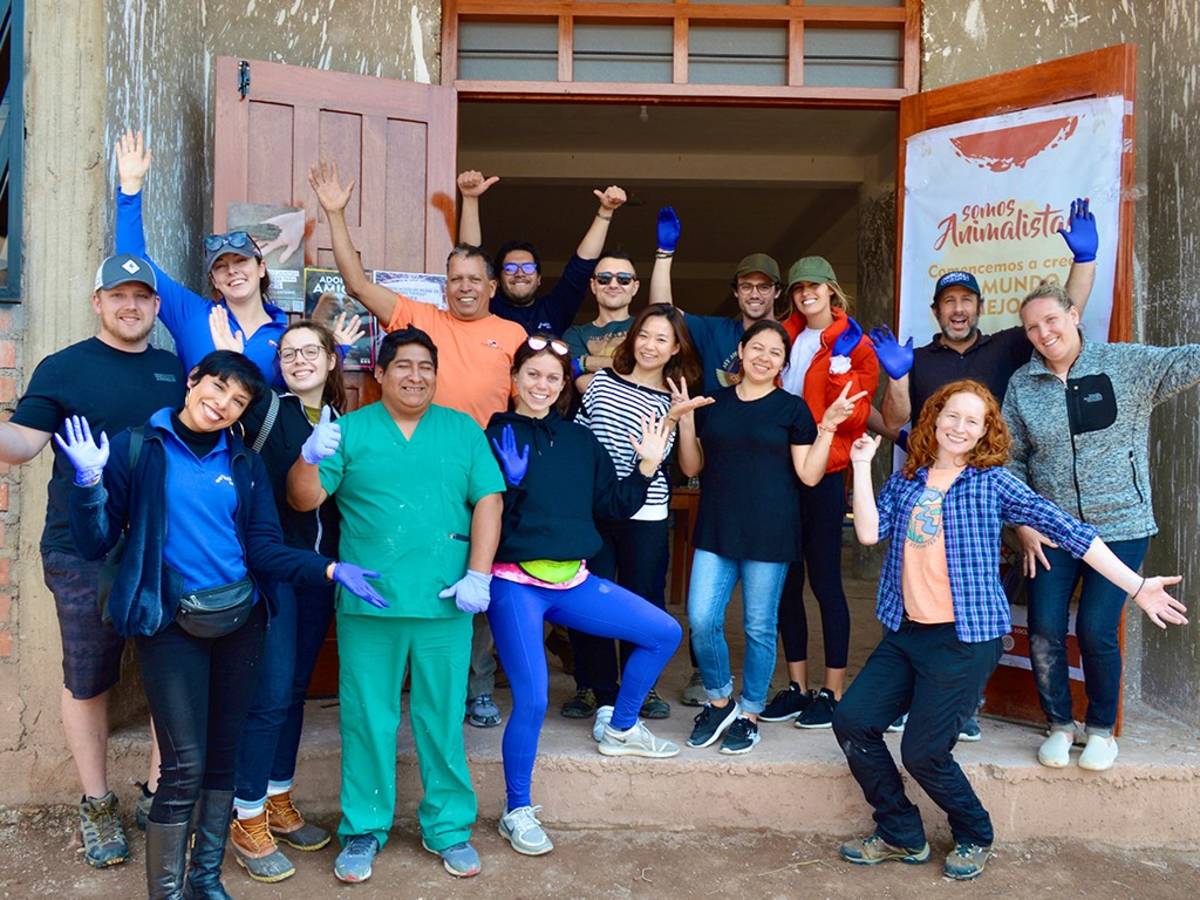 A diverse group of cheerful volunteers and staff pose together outside a community building in Peru, many wearing blue gloves and waving.