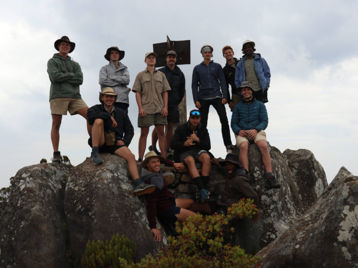 A group of eleven hikers pose on a rocky peak, dressed in outdoor gear with hats and jackets. The mood is cheerful, capturing a sense of adventure.