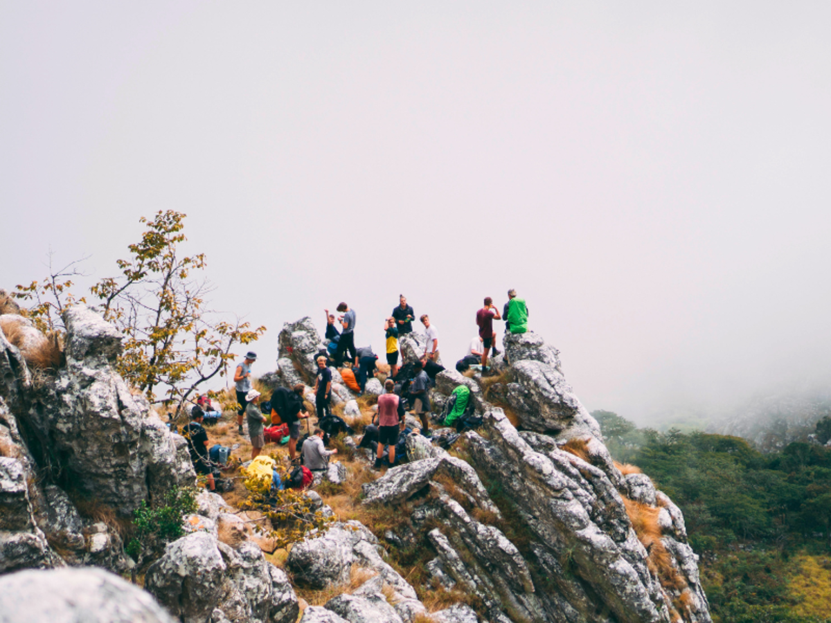 A group of hikers, wearing colorful clothing, gathers atop rocky cliffs surrounded by trees and mist, conveying a sense of adventure and camaraderie.