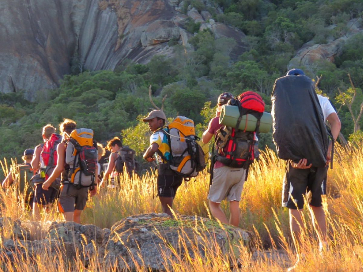 A group of hikers with backpacks walking through sunlit tall grass, surrounded by rocky cliffs and greenery, conveying adventure and camaraderie.