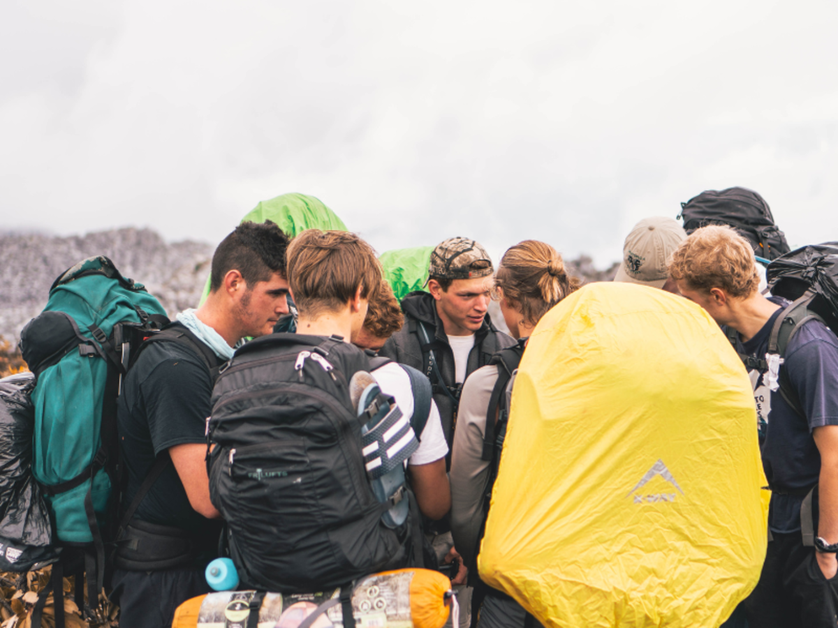A group of hikers with colorful backpacks huddle together, appearing focused and engaged in conversation against a cloudy mountain backdrop.