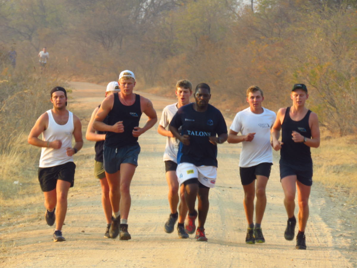 A group of seven men jogging on a dirt path surrounded by dry, sparse vegetation. They appear focused and determined. The mood is energetic and active.