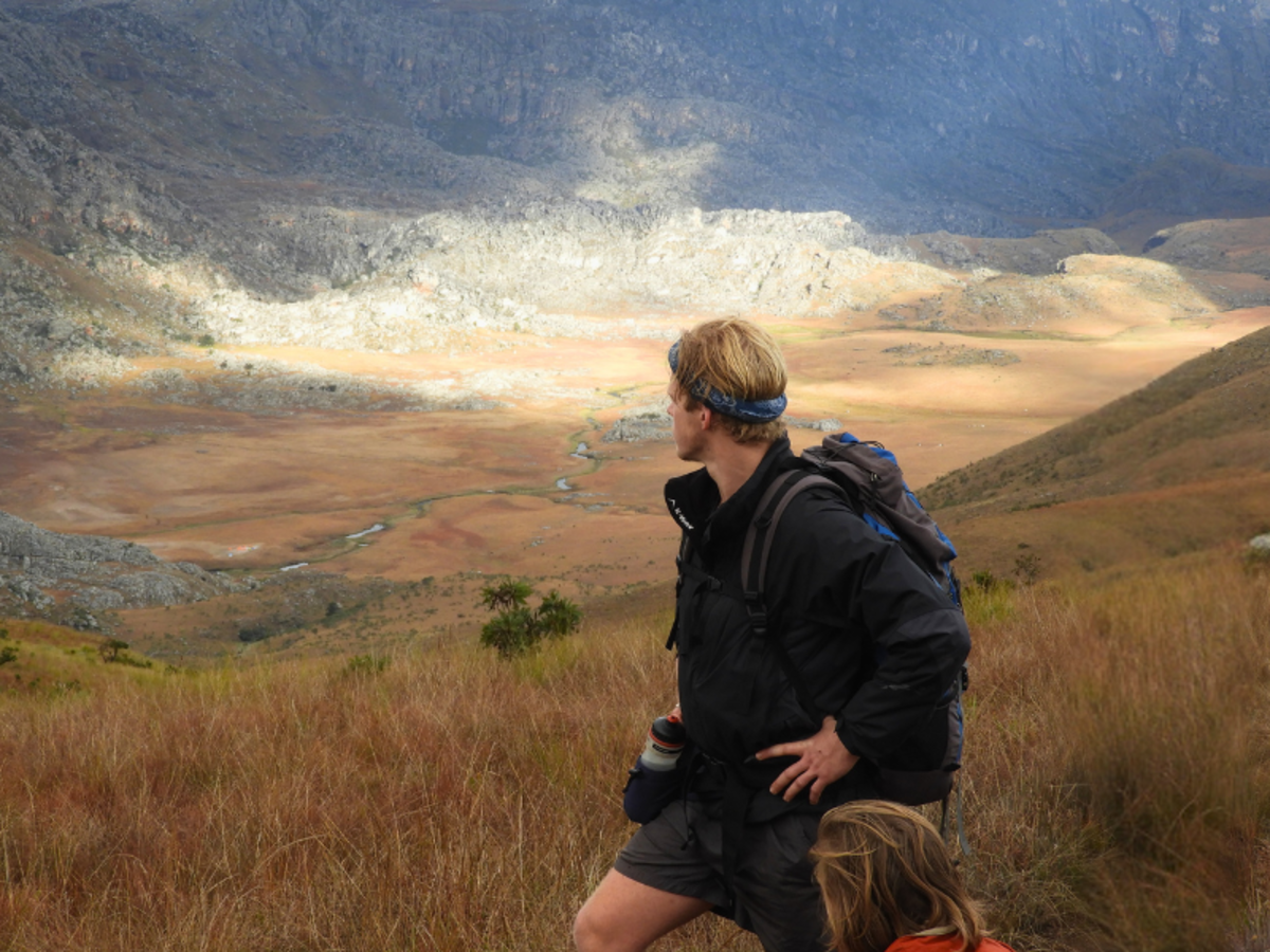 A hiker with a backpack stands on grassy terrain, looking over a vast, sunlit valley surrounded by rugged mountains, conveying a sense of adventure.