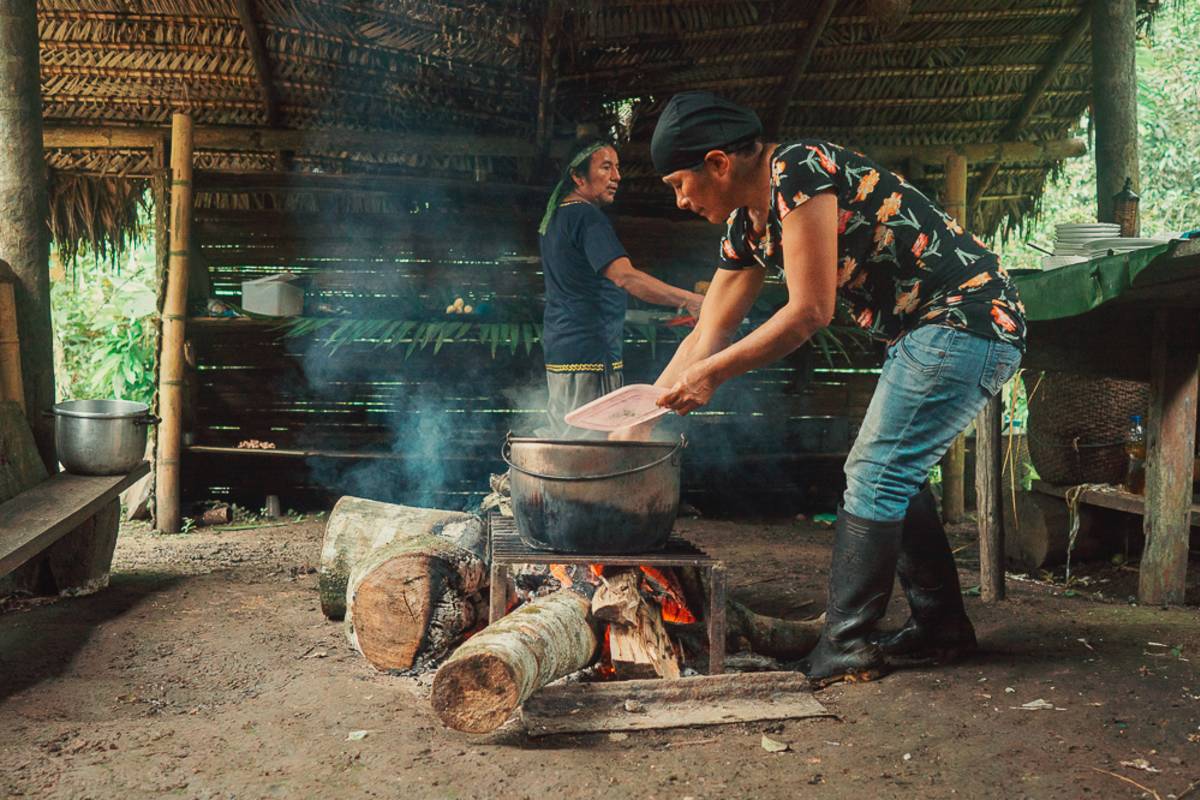 A local woman leans over pot cooking on the fire, in rustic outdoor kitchen.