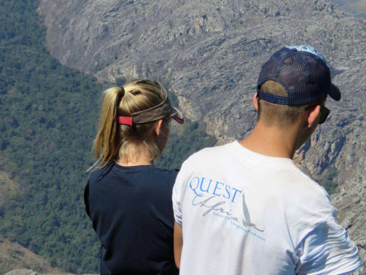 A man and woman, facing away, stand on a rocky viewpoint overlooking a forested valley. Both wear casual outdoor clothing, suggesting a hiking trip.