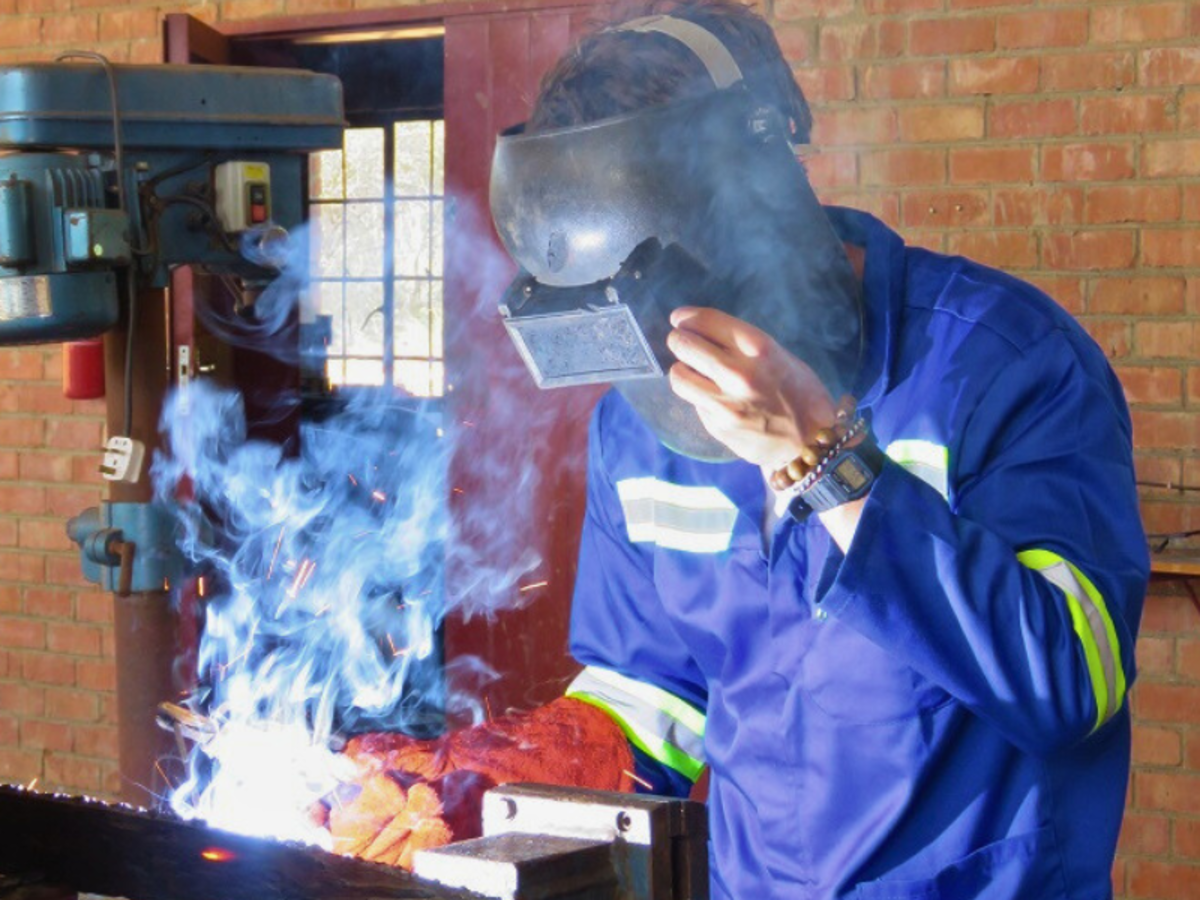 A person in a blue protective suit and welding helmet welds metal indoors, surrounded by blue smoke. The scene conveys focus and industry.