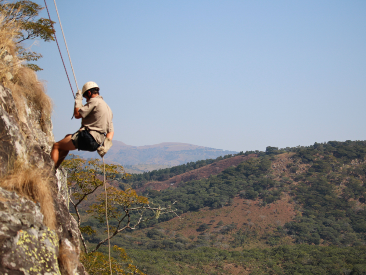 A person in a helmet rappels down a steep, rocky cliff against a backdrop of lush green hills and a clear blue sky, conveying adventure and excitement.